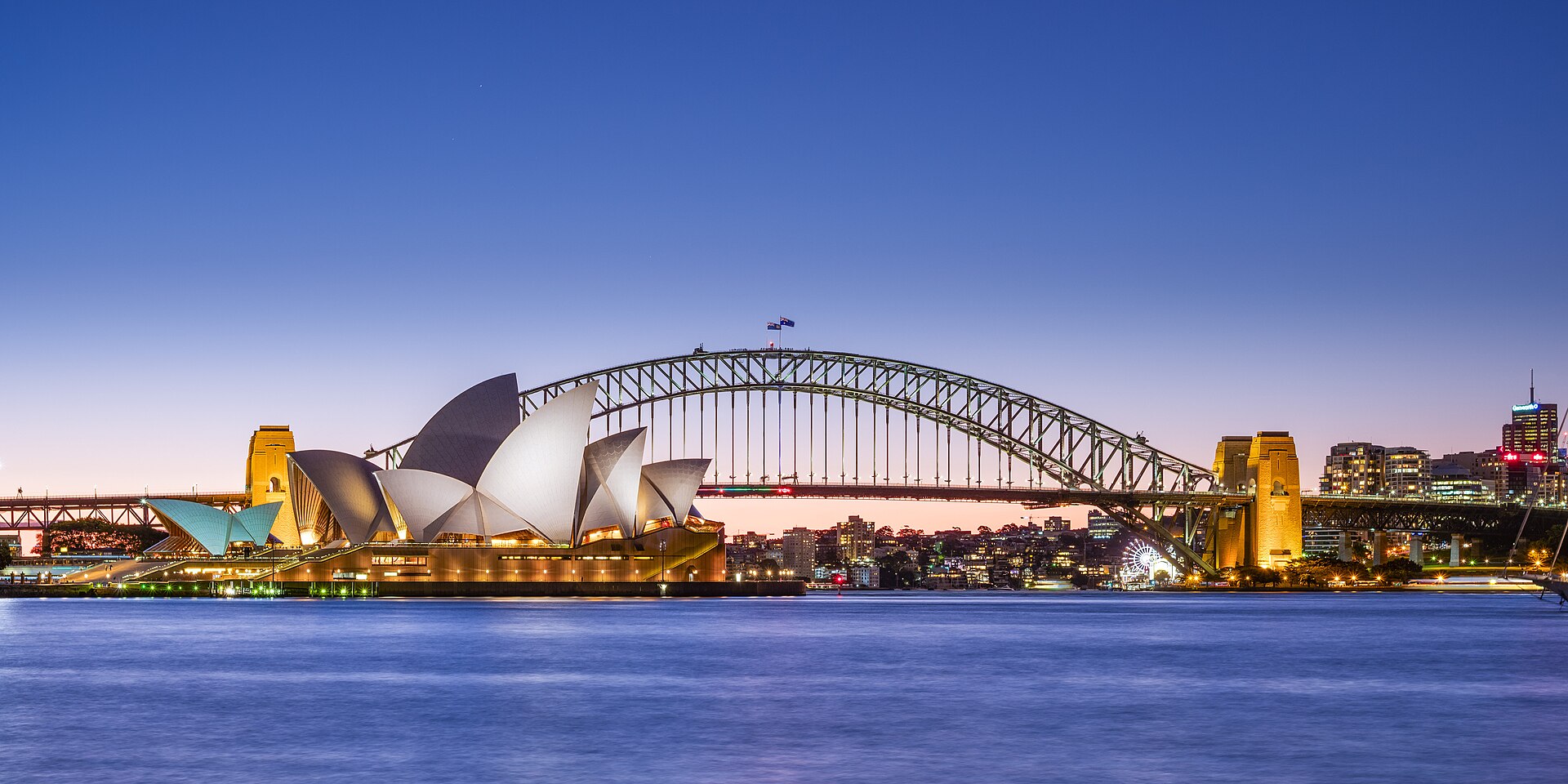 Jørn Utzon's Sydney Opera House, and the Harbour Bridge, two of Sydney's most famous landmarks, taken at late dusk. The Sydney Opera House is one of the most iconic buildings built in the 20th century (1973) and is UNESC